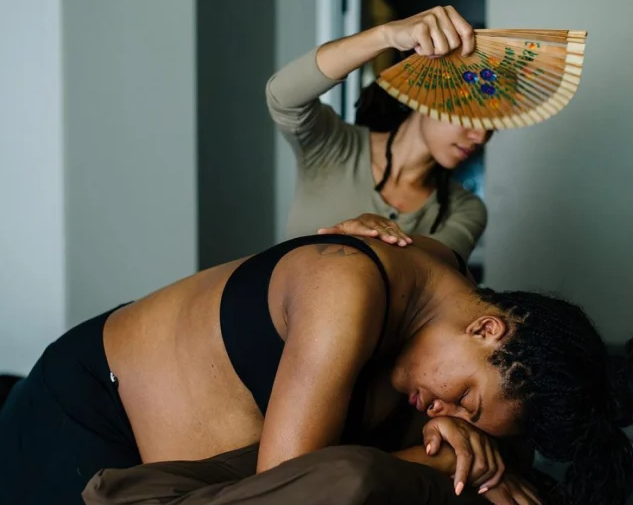 A pregnant woman leans over with her head resting on her hand as another woman fans her and puts a hand on her for support