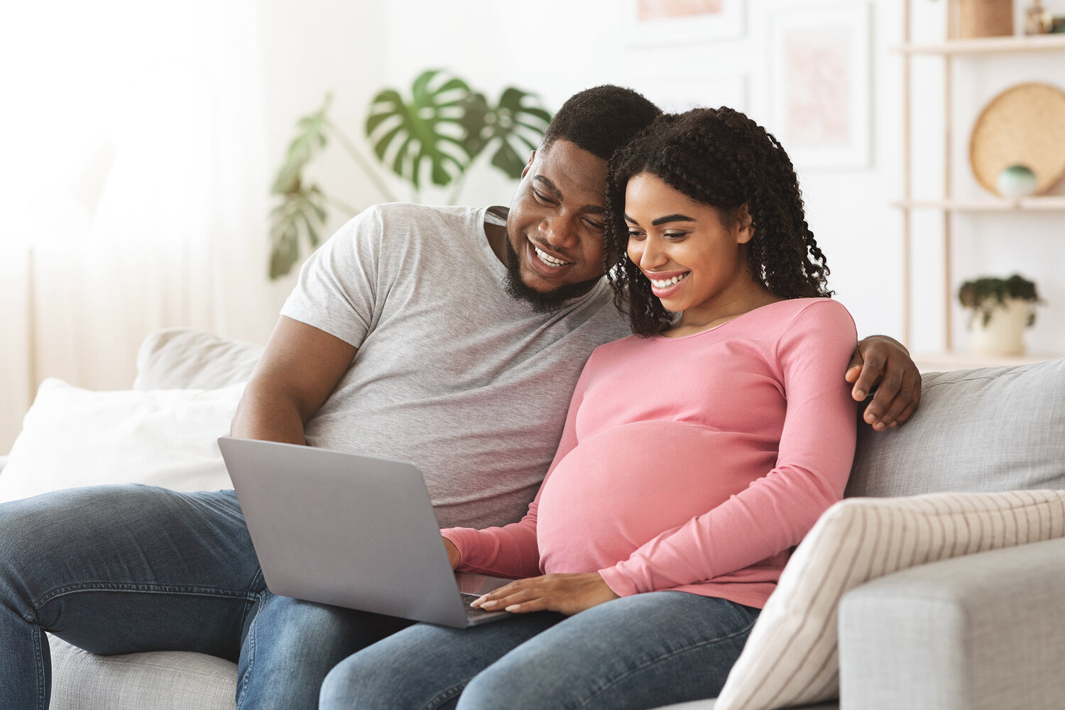 A pregnant woman and her partner sit on a sofa, viewing a laptop screen