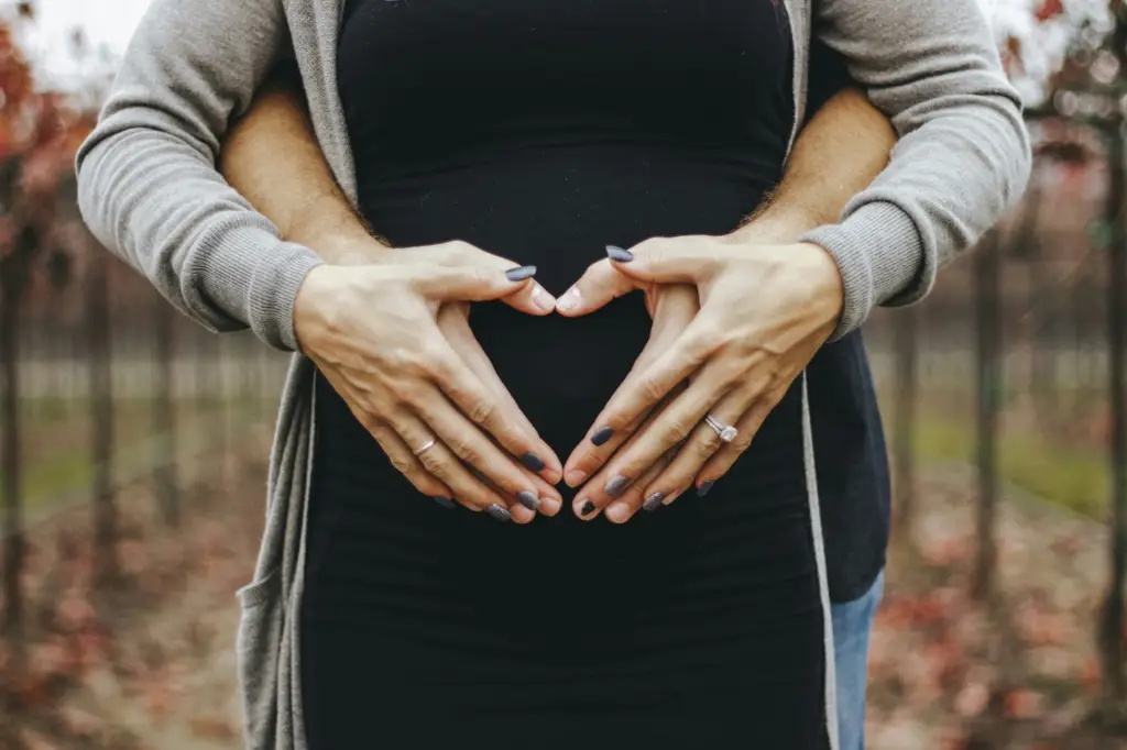 Man and woman embracing, forming a heart shape with their hands over her baby bump.